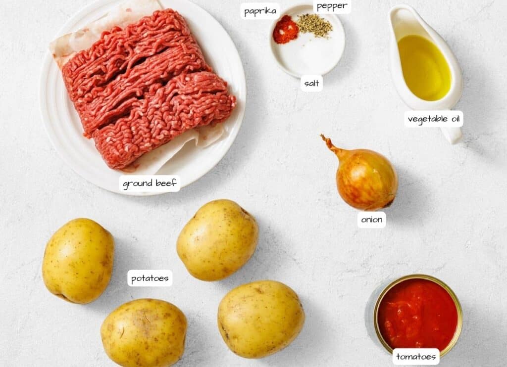 Overhead shot of labeled ingredients for "Poor Man's Meal": ground beef, potatoes, onion, canned tomatoes, vegetable oil, and spices (paprika, pepper, salt).