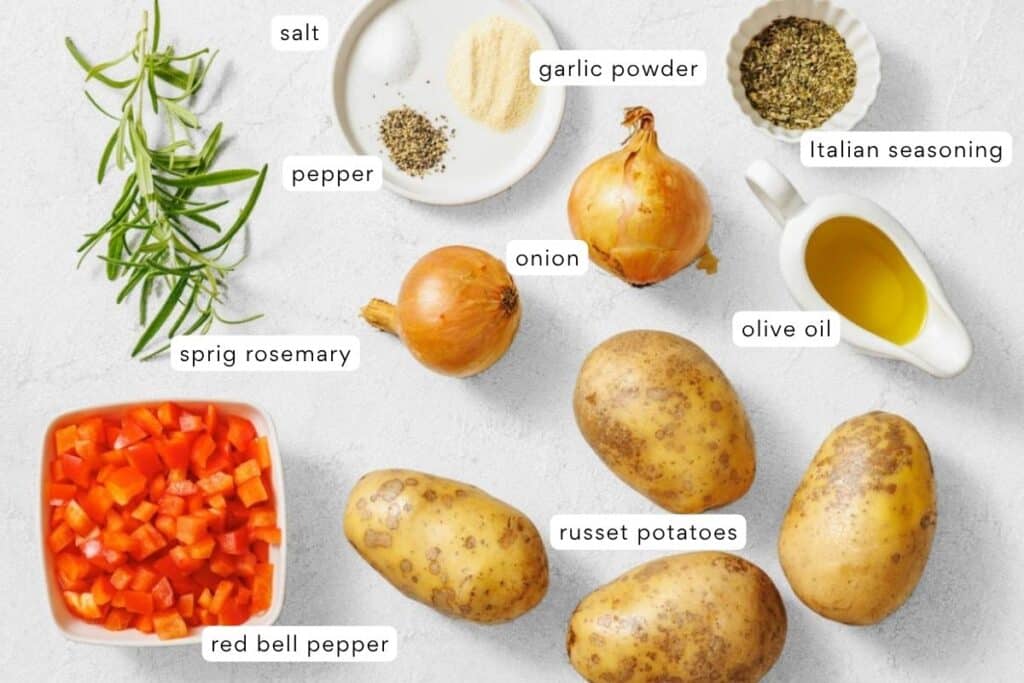 A selection of ingredients including russet potatoes, onions, red bell pepper, sprig rosemary, salt, pepper, garlic powder, Italian seasoning, and olive oil arranged on a white surface.
