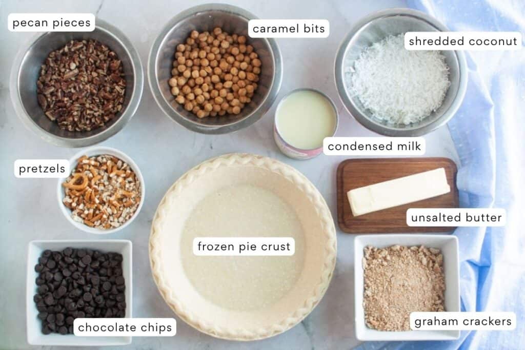 Top-down view of labeled baking ingredients including pecan pieces, caramel bits, shredded coconut, pretzels, condensed milk, unsalted butter, graham crackers, chocolate chips, and a frozen pie crust.