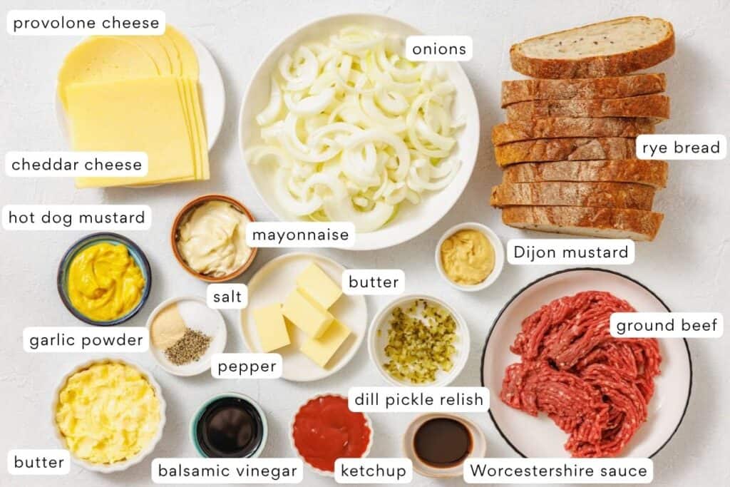 A variety of ingredients for making patty melts, including cheeses, sliced onions, rye bread, ground beef, sauces, seasonings, and condiments arranged on a white surface.