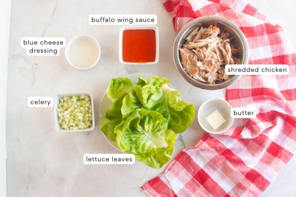 Ingredients for buffalo chicken lettuce wraps arranged on a counter: blue cheese dressing, celery, lettuce leaves, buffalo wing sauce, shredded chicken, and butter.