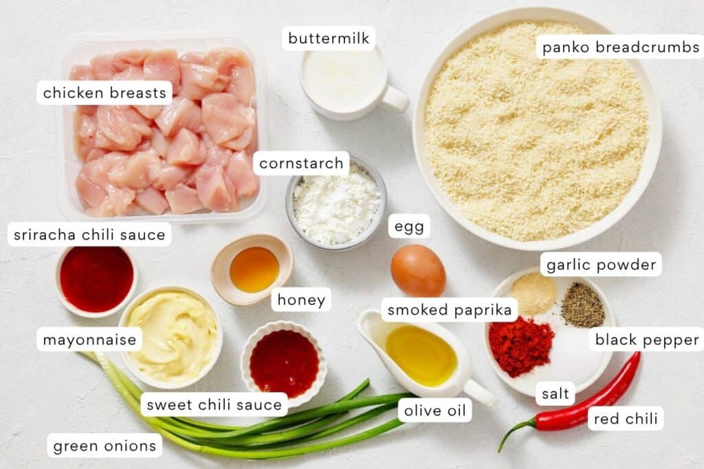 Various ingredients arranged on a white surface, including diced chicken breasts, seasonings, sauces, egg, buttermilk, panko breadcrumbs, and green onions, each labeled with text.
