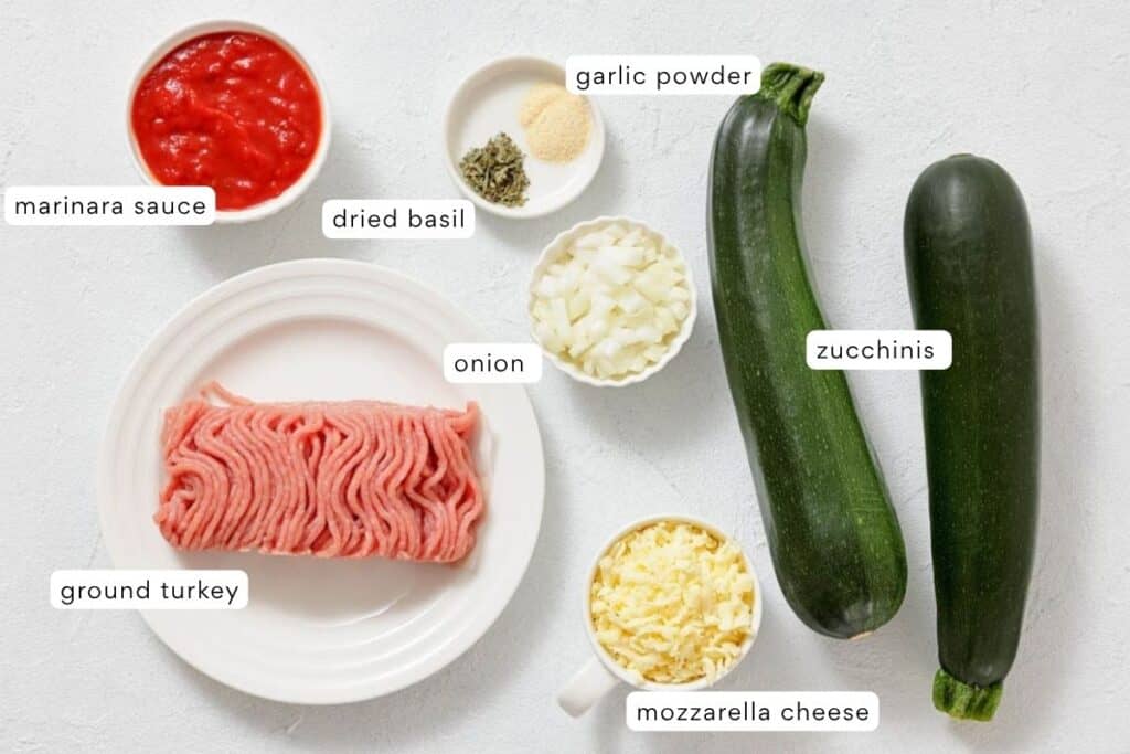 Ingredients for a dish laid out on a white surface: marinara sauce, ground turkey, dried basil, garlic powder, chopped onion, zucchinis, and shredded mozzarella cheese.