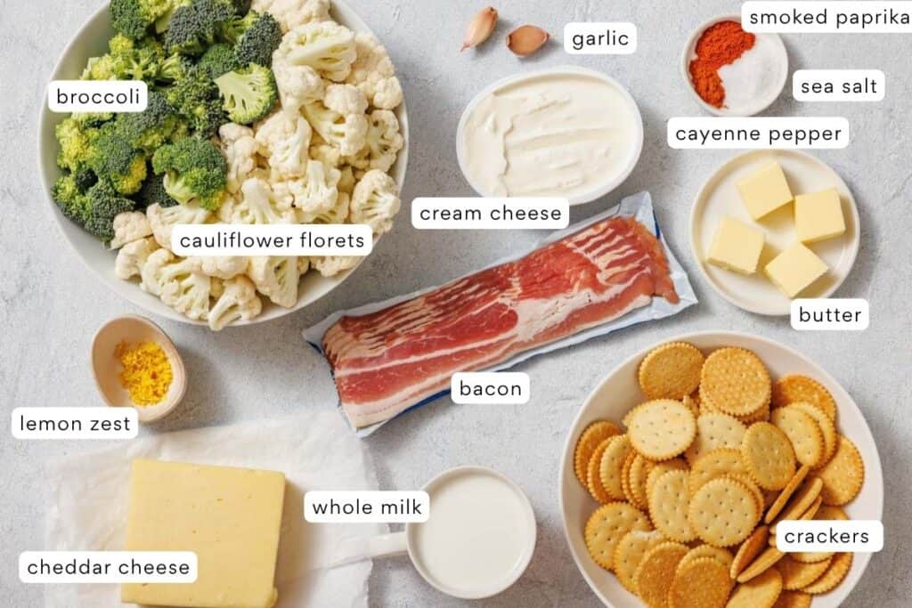 Various ingredients are arranged on a table, including broccoli, cauliflower florets, cream cheese, bacon, cheddar cheese, milk, crackers, butter, garlic, lemon zest, and spices.