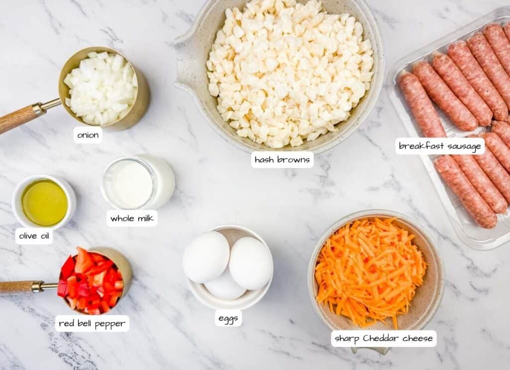Ingredients for a breakfast casserole laid out on a marble surface, including chopped onion, olive oil, red bell pepper, whole milk, eggs, hash browns, sharp cheddar cheese, and breakfast sausage.