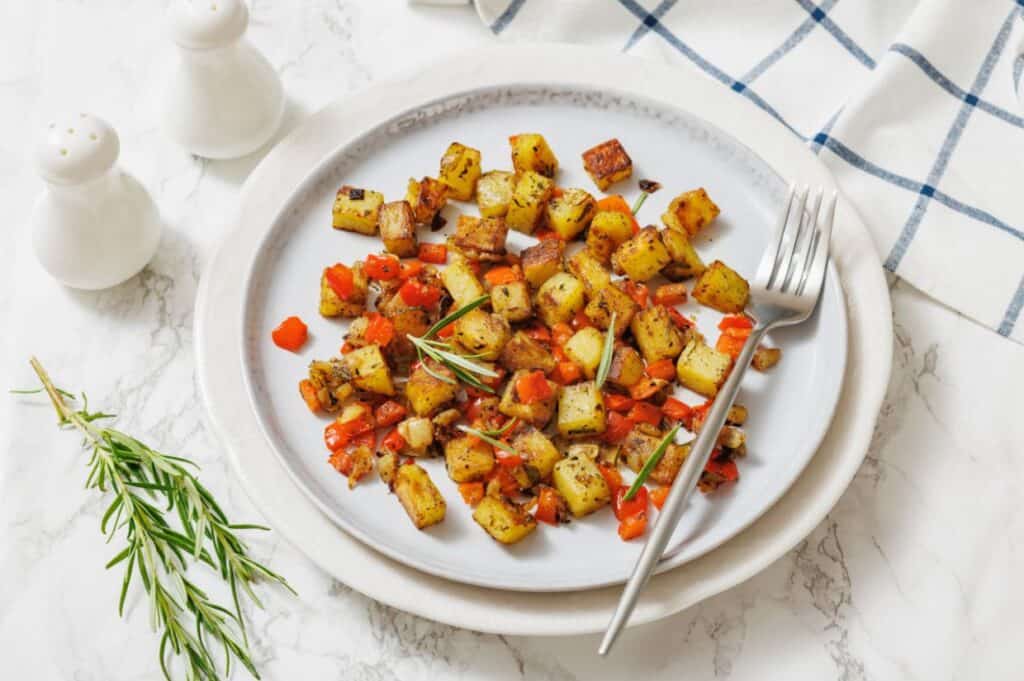 A white plate with diced, cooked potatoes, red bell peppers, and herbs, placed on a marble surface with a fork, fresh rosemary, and salt and pepper shakers nearby.