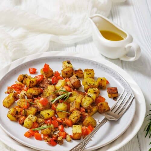 A plate of seasoned roasted potatoes and red bell peppers with a fork, garnished with herbs, sits next to a small sauce pitcher of olive oil on a white table.