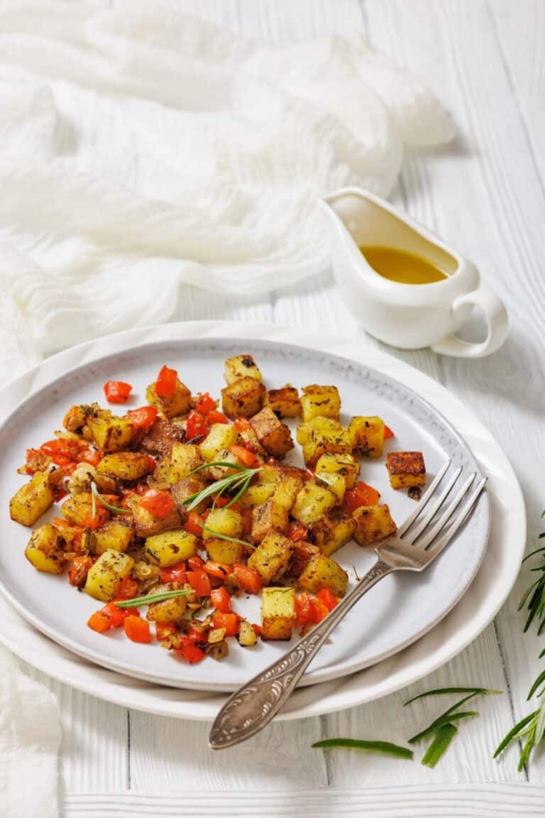 A plate of seasoned roasted potatoes and red bell peppers with a fork, garnished with herbs, sits next to a small sauce pitcher of olive oil on a white table.