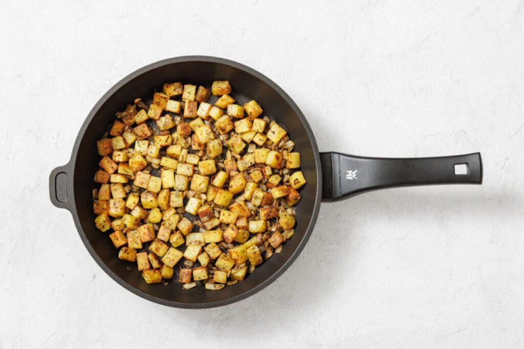 A black frying pan filled with evenly diced, seasoned, and cooked potatoes on a white surface.