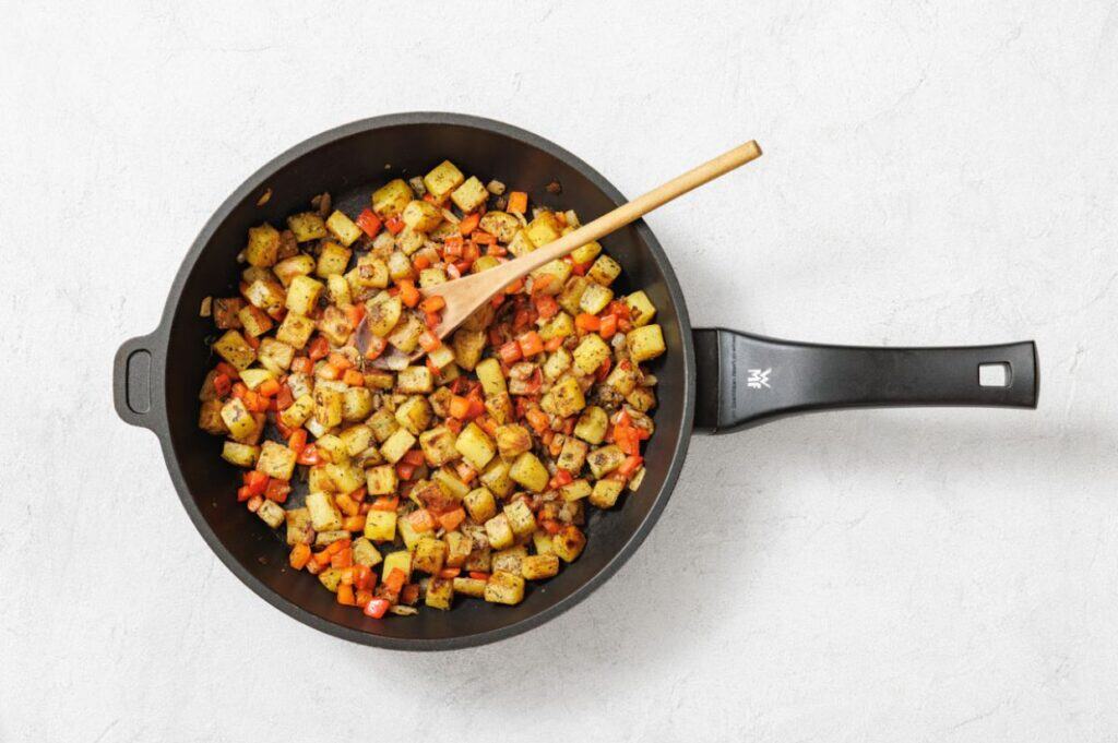 A frying pan with sautéed diced potatoes, carrots, onions, and spices, with a wooden spoon resting inside on a white background.