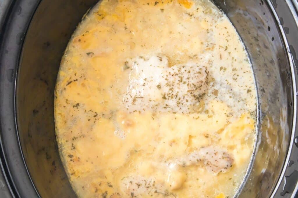 Chicken breasts covered in a creamy, seasoned sauce are cooking inside a slow cooker.