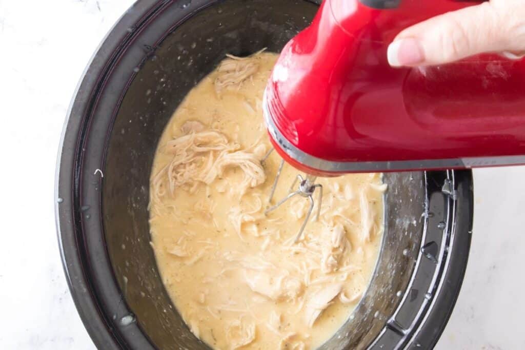 A hand uses a red electric hand mixer to shred cooked chicken in a creamy sauce inside a black slow cooker.