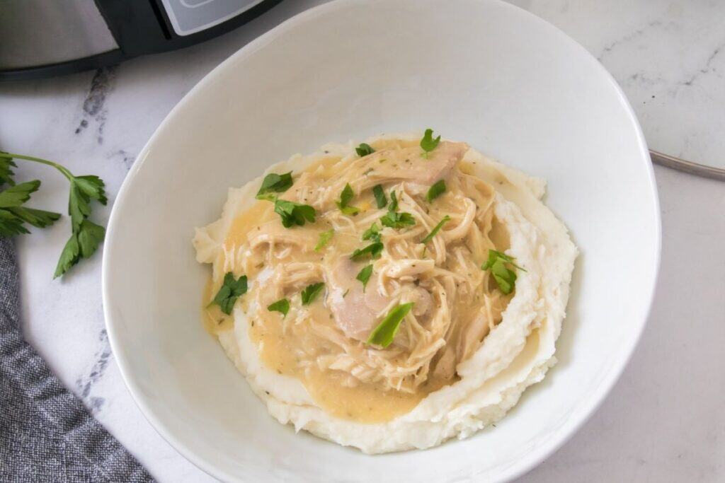 A bowl of mashed potatoes topped with shredded chicken and creamy sauce, garnished with fresh parsley.