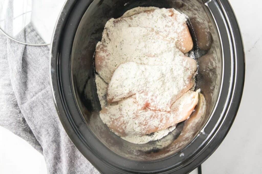 Raw chicken breasts covered with a layer of flour in a slow cooker, with a gray kitchen towel and the glass lid placed to the side.
