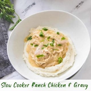 A slightly high-angle shot of a white bowl filled with creamy white mashed potatoes, topped with shredded "Slow Cooker Ranch Chicken & Gravy" and fresh green parsley garnish, on a white marble surface.