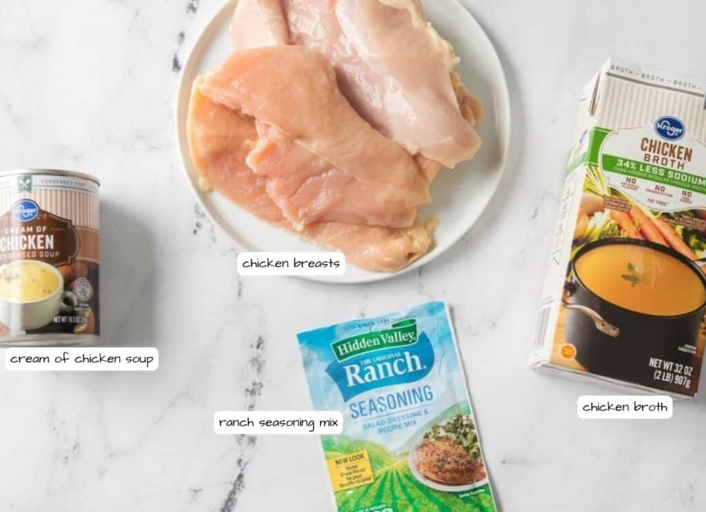 Raw chicken breasts on a plate, a can of cream of chicken soup, a packet of ranch seasoning mix, and a carton of chicken broth arranged on a white surface.