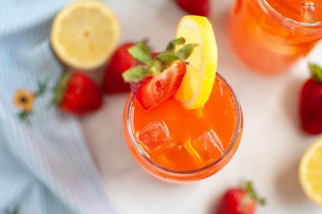 An overhead shot of a clear glass of bright orange-red strawberry lemonade with ice cubes, garnished with a halved fresh strawberry and a yellow lemon slice on the rim, with other fruit and a blue and white striped napkin in the background.