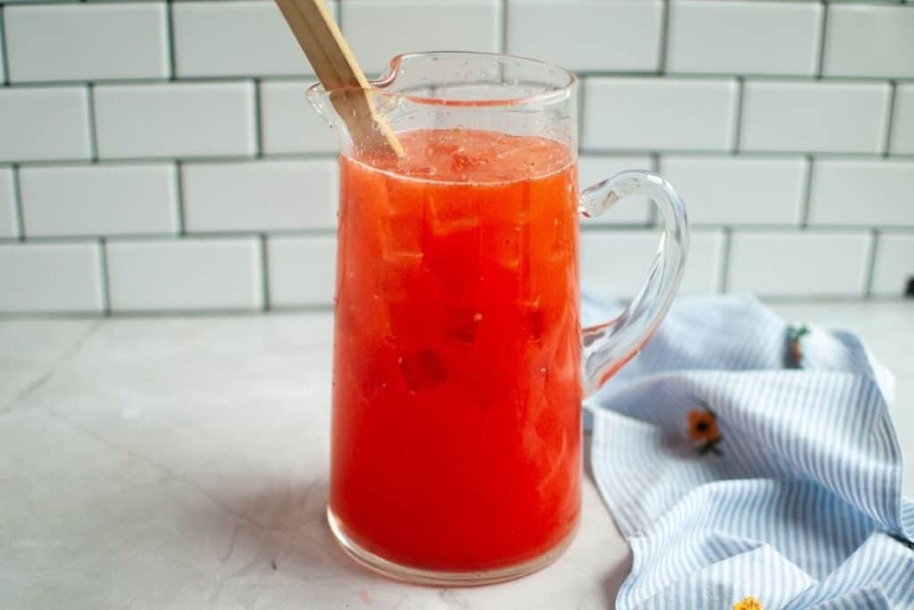A clear glass pitcher, with a handle on the right, is filled with bright red strawberry lemonade and ice cubes, with a light wooden stirring spoon resting inside, on a white countertop with a blue and white striped napkin and a white tiled backsplash.