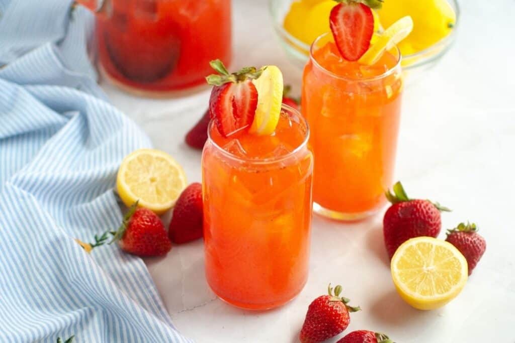 Two clear glasses of bright red strawberry lemonade with ice, garnished with halved strawberries and lemon slices, with whole strawberries and lemon slices scattered around them on a white surface, and a blurred pitcher in the background.