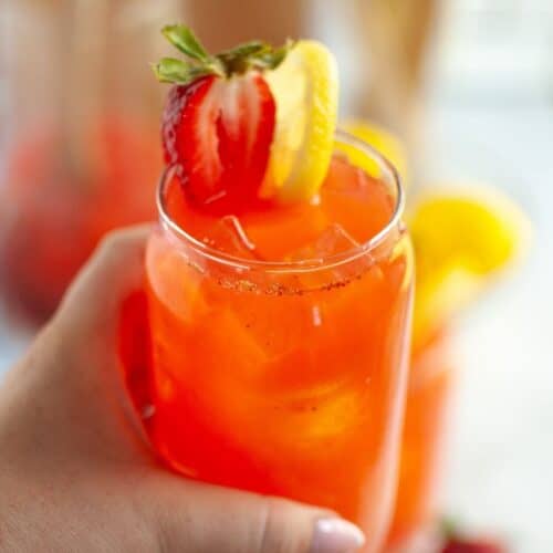 A close-up of a hand with painted fingernails holding a clear glass filled with bright red strawberry lemonade and ice, garnished with a halved strawberry and a lemon slice on the rim, with blurred drinks in the background.