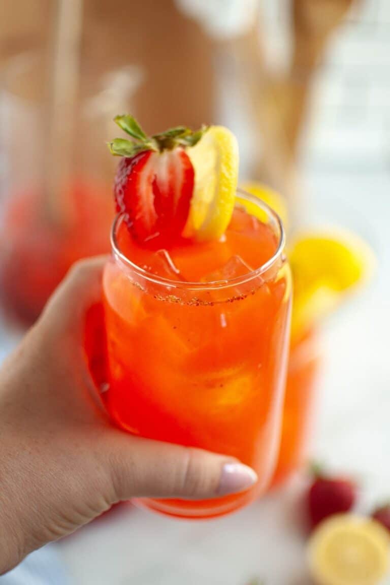 A close-up of a hand with painted fingernails holding a clear glass filled with bright red strawberry lemonade and ice, garnished with a halved strawberry and a lemon slice on the rim, with blurred drinks in the background.