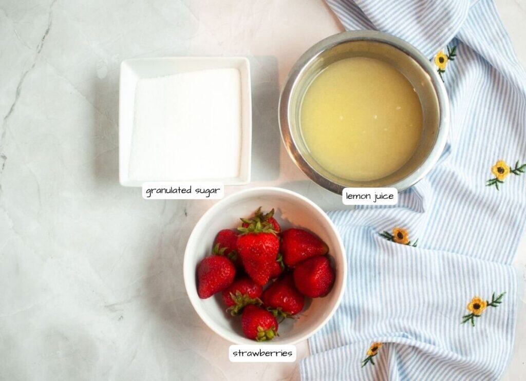 An overhead view of a white countertop displaying labeled ingredients for Strawberry Lemonade: a square dish of granulated sugar, a metal bowl of lemon juice, and a white bowl of whole red strawberries, with a blue and white striped napkin.