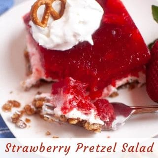 A slice of strawberry pretzel salad with whipped cream and a pretzel on top, served on a white plate with a strawberry and a fork.