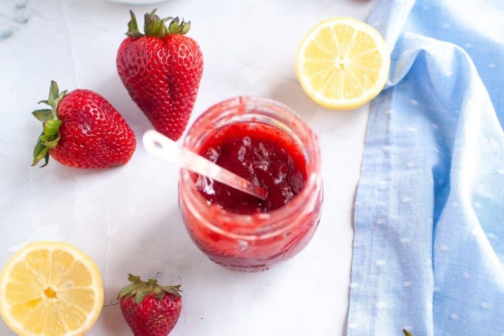 Overhead shot of a clear glass jar filled with vibrant red strawberry puree and a plastic spoon, surrounded by fresh whole strawberries and lemon halves on a white surface with a blue cloth.