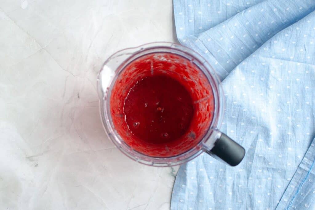 Overhead shot of a blender jar containing a vibrant red, slightly chunky strawberry puree, with a blue cloth nearby.