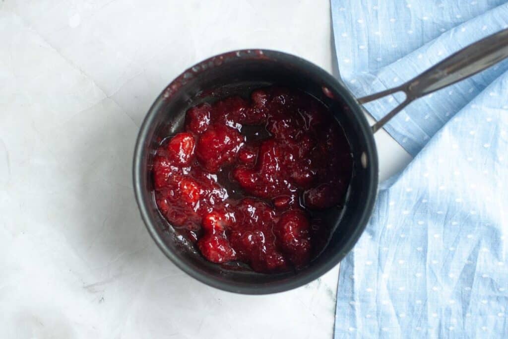 Overhead shot of a dark nonstick saucepan filled with glossy, deep red cooked strawberries, some whole and some broken down.