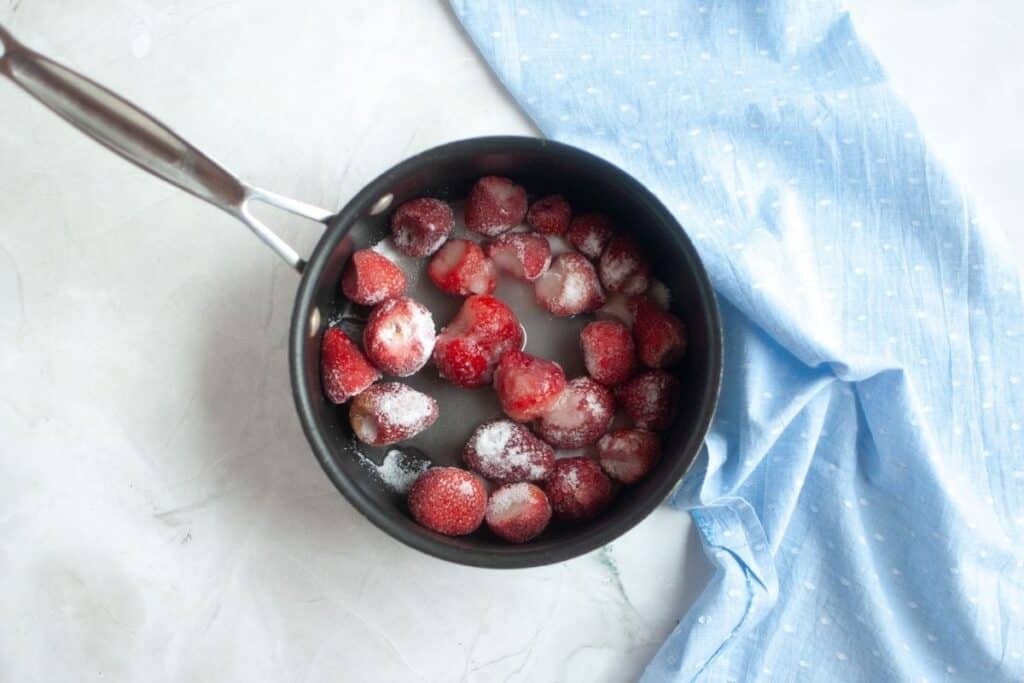 Overhead shot of a dark nonstick saucepan filled with red strawberries, many coated in white sugar crystals, with a blue cloth nearby.