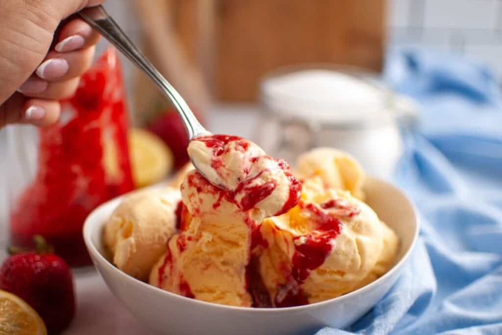 A hand holds a spoonful of red strawberry puree over scoops of vanilla ice cream in a white bowl, with fresh strawberries and a lemon in the background.