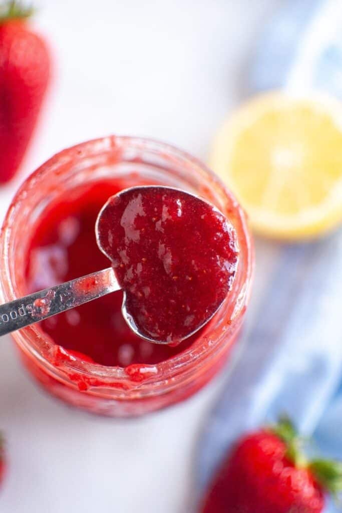 A metal spoon, shaped like a heart, holds a spoonful of vibrant red strawberry puree above a glass jar, with fresh strawberries and a lemon in the background.