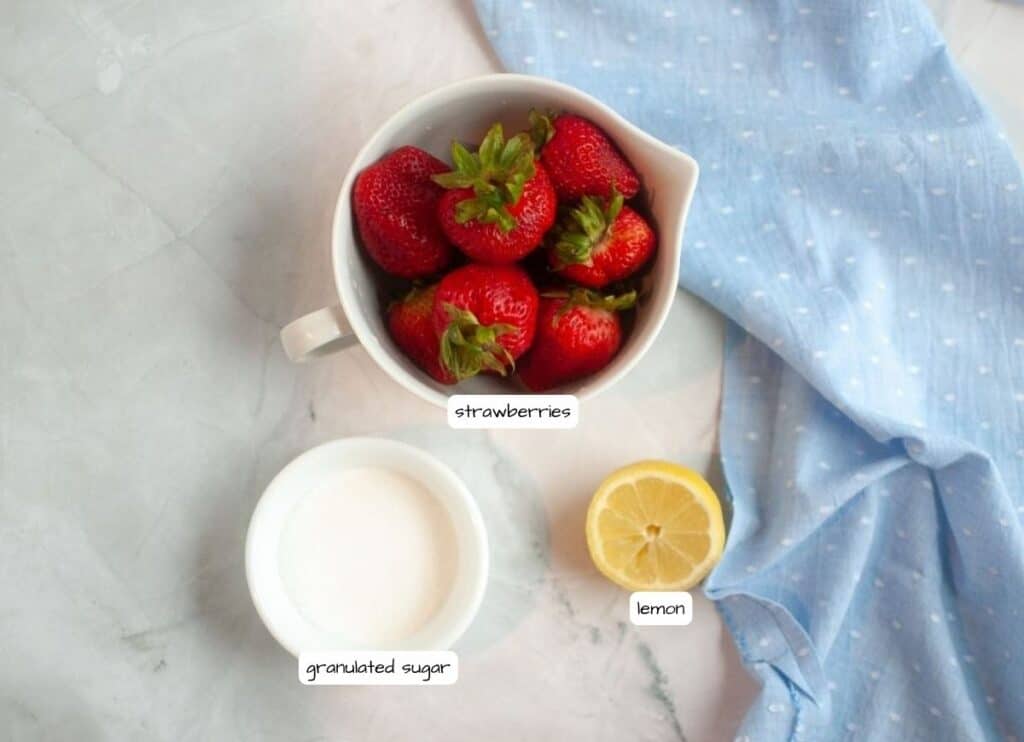 Overhead shot of labeled ingredients for strawberry puree: fresh strawberries in a white pitcher, a small bowl of granulated sugar, and a lemon half, on a marble surface with a blue cloth.