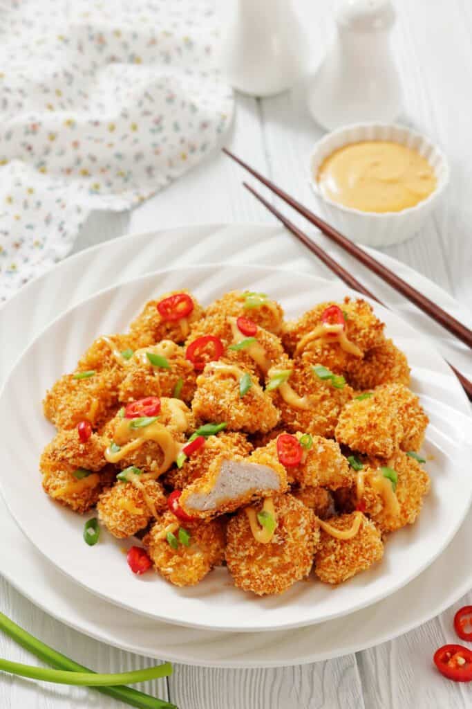 A plate of breaded chicken nuggets garnished with sliced red chili, green onions, and drizzled sauce, with chopsticks and a dipping sauce on the side.