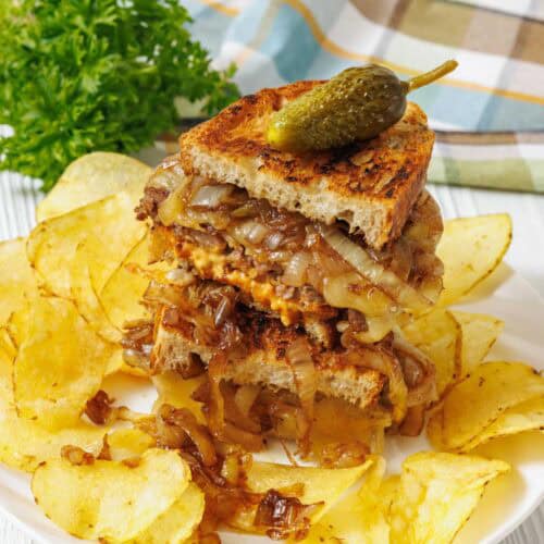 A plate with a stacked grilled sandwich filled with caramelized onions and meat, topped with a pickle, surrounded by potato chips. A bunch of parsley and a checkered cloth in the background.