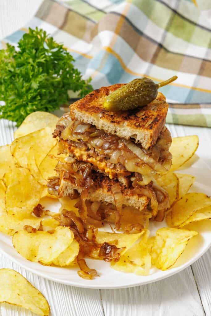 A plate with a stacked grilled sandwich filled with caramelized onions and meat, topped with a pickle, surrounded by potato chips. A bunch of parsley and a checkered cloth in the background.