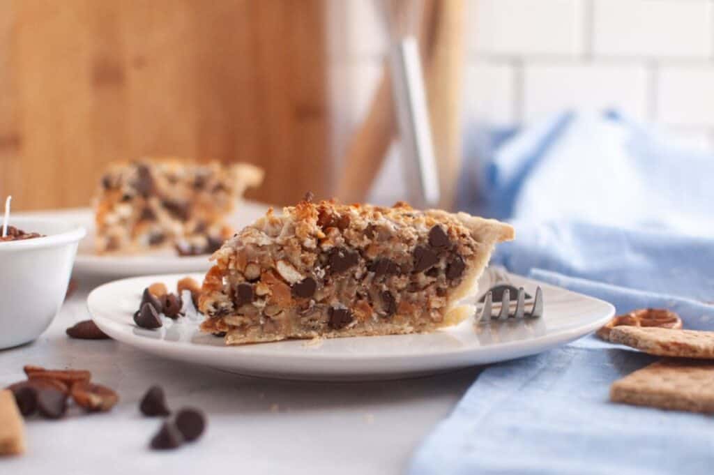 A slice of pie filled with chocolate chips, nuts, and coconut sits on a white plate with a fork, with a bowl and another slice in the background.