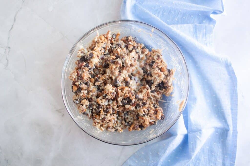 A glass bowl filled with a mixture of oats, raisins, and nuts sits on a light marble surface beside a folded blue cloth.