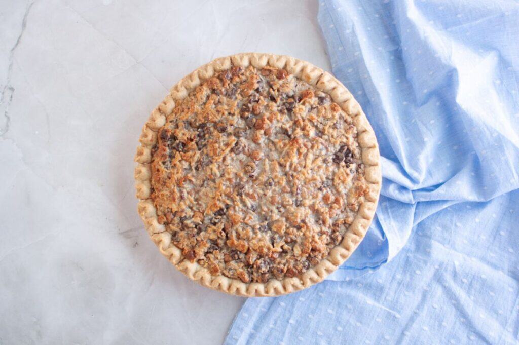 A whole baked pie with a crumbly topping sits on a light marble surface next to a light blue, textured cloth.