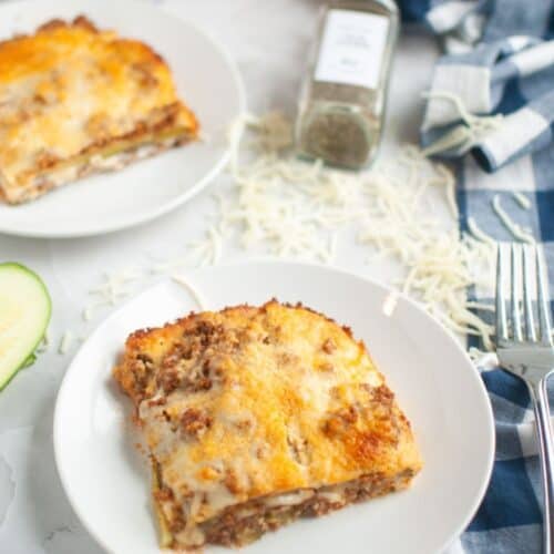 Two slices of zucchini lasagna are served on white plates, with shredded cheese, a spice jar, and a blue checkered napkin in the background.