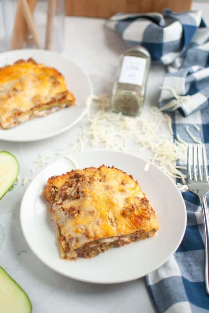 Two slices of zucchini lasagna are served on white plates, with shredded cheese, a spice jar, and a blue checkered napkin in the background.