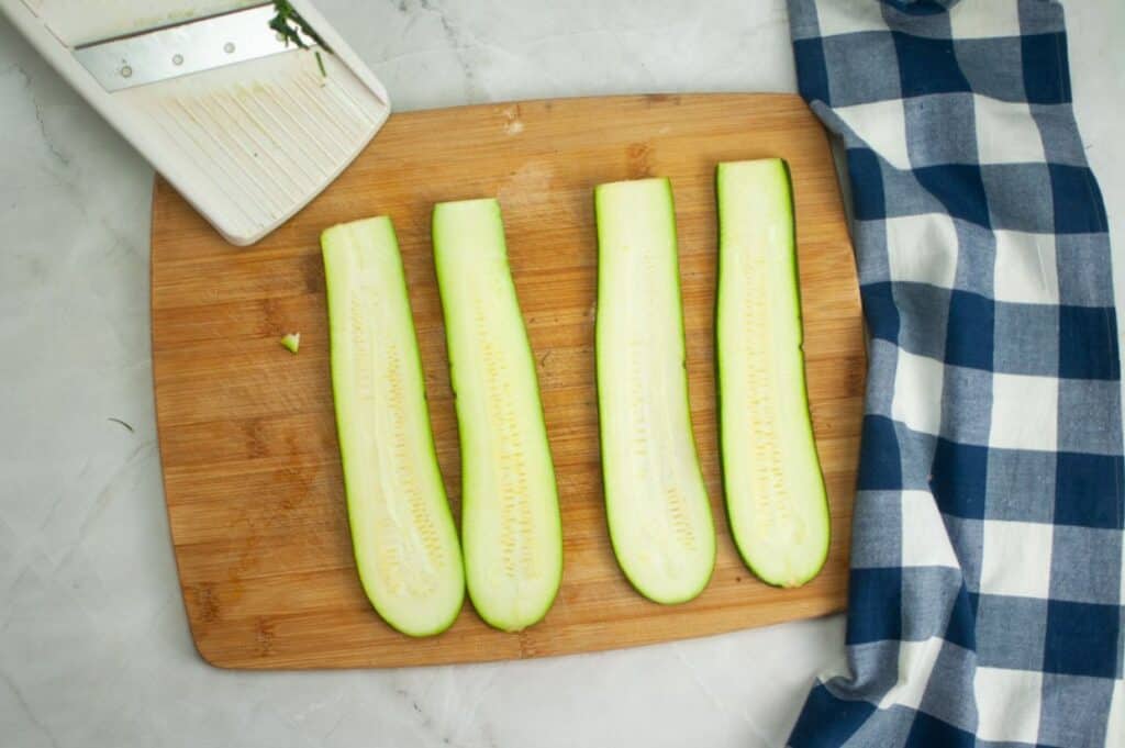 An overhead view of a wooden cutting board with four long, light green zucchinis sliced lengthwise, revealing their pale green flesh and small white seeds, with a white mandoline slicer in the upper left corner.