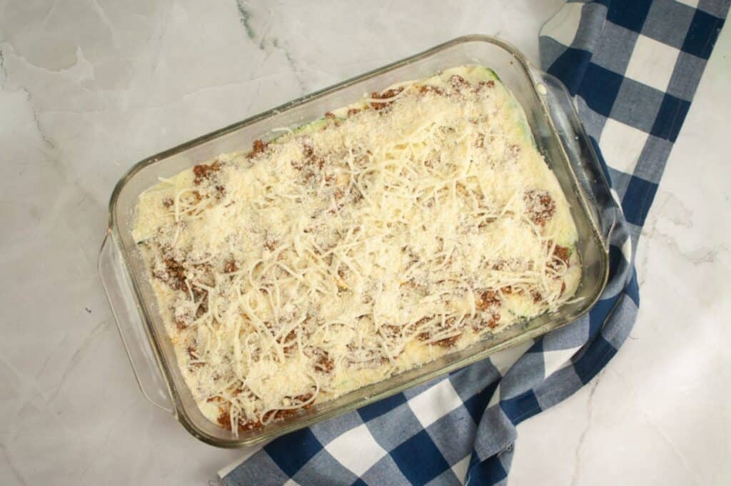 An overhead view of a rectangular clear glass baking dish filled with an assembled, uncooked zucchini lasagna, generously covered with a layer of white shredded cheese and some smaller grated cheese, with a blue and white checkered napkin to the right.