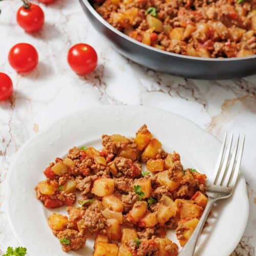 A white plate holds a serving of Poor Man's Meal (ground meat and potato hash) garnished with parsley, with a skillet of the dish and cherry tomatoes in the background.