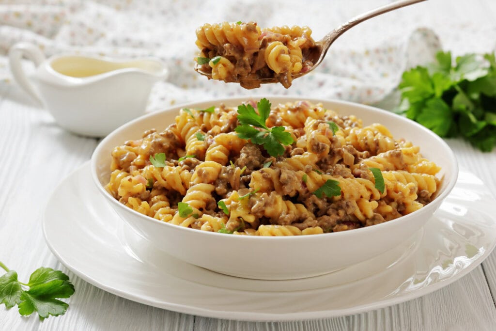 A bowl of cooked rotini pasta mixed with ground meat and sauce, garnished with parsley, with a spoon lifting a portion above the bowl.