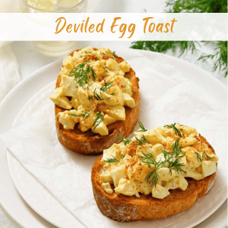 Two slices of toasted bread topped with chopped deviled eggs and garnished with fresh dill are served on a white plate.