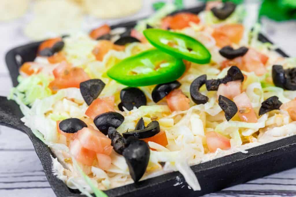 A black dish contains a layered dip topped with shredded lettuce, diced tomatoes, sliced black olives, and two slices of jalapeño pepper.