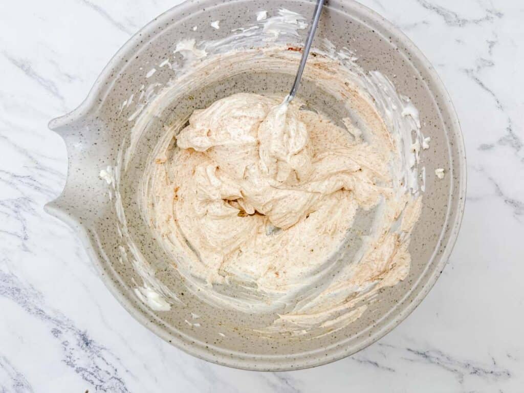 A mixing bowl containing a light brown, whipped batter with a spoon inside, placed on a marble countertop.