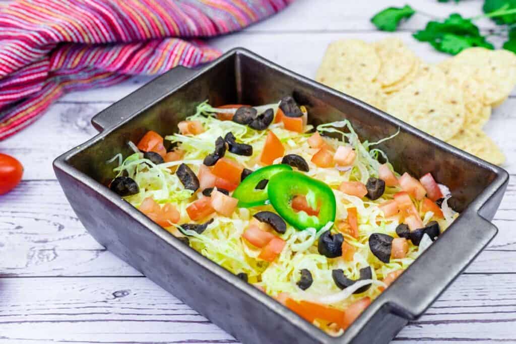 Rectangular dish filled with layered taco dip topped with shredded lettuce, diced tomatoes, black olives, onions, and jalapeño slices. Tortilla chips and a striped cloth are nearby.